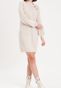 Robe pull en tricot beige clair avec un col monté, des manches longues bouffantes et trois boutons couleur or sur l'épaule. Coupe ajustée.