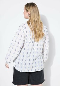 Vit långärmad blus med blå geometriska mönster och lätt tyg. Matchad med mörka shorts som har rundad nederkant och en lös passform.