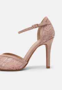 Chaussure à talon haut rose blush avec bride à la cheville, superposition en dentelle florale sur le devant et le talon, et talon aiguilles fin sur fond blanc.