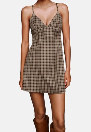 Mujer con un mini vestido marrón a cuadros sin mangas con tirantes finos y un escote en V profundo, combinado con botas altas marrones.