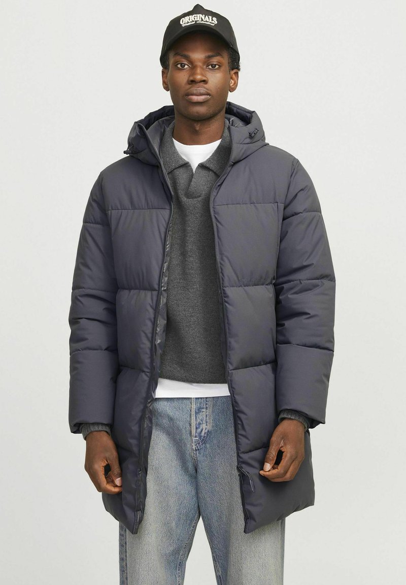 Marineblå pufferjakke med hætte, quiltet design og frontlommer, båret over en grå sweater. Sammen med lyseblå denimjeans.