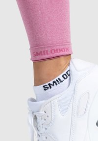 Vaaleanpunaiset urheilulegginsit, joissa on kääritty lahje ja "SMILODOX"-logo, yhdistetty valkoisiin tennareihin ja mustaruudullisiin sukkiin.