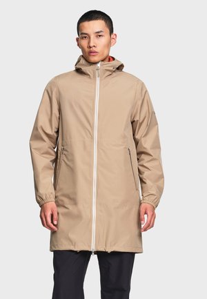 Homme portant un long imperméable beige à capuche avec fermeture éclair à l'avant et poches latérales zippées, debout devant un fond uni.