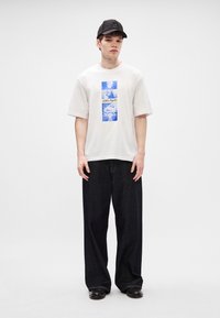 Λευκό oversized t-shirt με μπλε και λευκό γραφικό σχέδιο στο κέντρο, σε συνδυασμό με χαλαρές μαύρες τζιν παντελόνες και μαύρο καπέλο.