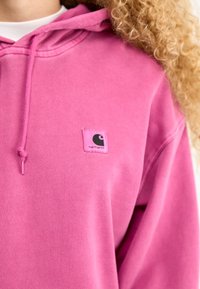 Hoodie rosa con un cordino, caratterizzato da una piccola patch quadrata con il logo Carhartt nell'angolo in alto a sinistra. Materiale morbido in cotone con una texture liscia.