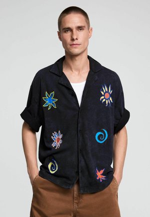 Junger Mann mit kurzen Haaren, der ein schwarzes Hemd mit bunten abstrakten Sonne- und Wirbelmustern trägt, die Hände in den Taschen einer braunen Hose.