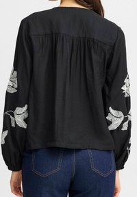 Blouse noire avec de longues manches bouffantes ornées de motifs de feuilles brodés en blanc. Tissu doux, dos froncé et coupe décontractée.