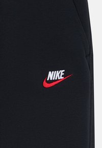 Svarta joggers i tyg med en broderad Nike-logotyp i vitt och rött. Slät textur med kontrasterande färgdetaljer.