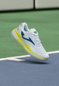Weißer Sportschuh mit blauen und gelben Akzenten, strukturiertem Mesh-Obermaterial, flexiblem Sohle und verstärkter Zehenkappe, entworfen für sportliche Nutzung.
