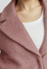 Gros plan sur une personne portant un manteau texturé rose poussiéreux par-dessus un haut blanc à col rond, montrant le col et le haut de la poitrine.