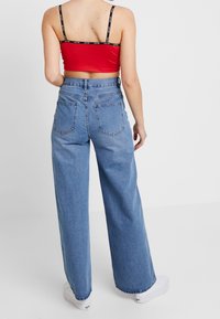 Jean bleu à jambes larges en denim, doté d'une taille haute et de deux poches arrière. La tenue comprend un top rouge court avec des bretelles noires.