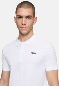Valkoinen polo-paita, joka on valmistettu puuvillasta. Siinä on klassinen kaulus, lyhyet hihat ja rintaosassa pieni punainen ja sininen FILA-logo.