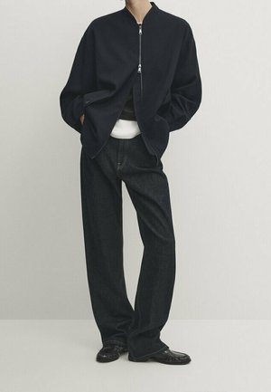 Person wearing a black bomber jacket, white shirt underneath, loose-fitting dark jeans, and black textured shoes, standing against a plain backdrop.