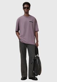 Camiseta morada de gran tamaño con un pequeño gráfico en el frente, combinada con jeans negros holgados y un bolso de cuero negro. Lleva gafas de sol negras.