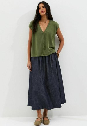 Kobieta ubrana w oliwkowozielony bezrękawnik na guziki z dekoltem w serek, ciemnoniebieską długą spódnicę oraz beżowe buty typu Mary Jane, stojąca na tle białej, gładkiej ściany.