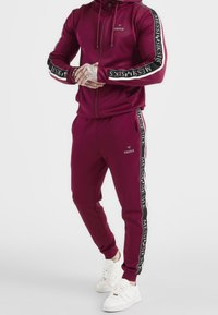 Mörkröd träningsoverall med zip-up jacka och matchande byxor. Funktioner logotypdetaljer i svart och vitt på ärmarna och sidosträck. Vita sneakers.