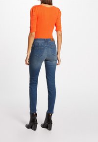 Kvinde, der vender væk, iført en lys orange kortærmet top, blå skinny jeans og sorte hælede ankelstøvler på ensfarvet hvid baggrund.