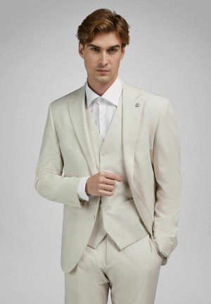 Junger Mann mit hellbraunen Haaren, gekleidet in einen cremefarbenen Dreiteiler-Anzug und weißem Hemd, eine Hand justiert die Jacke, neutraler grauer Hintergrund.