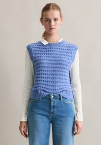 Teksturowana jasnoniebieska dzianinowa kamizelka z prążkowanym wykończeniem noszona na białej koszuli z długim rękawem. Modelka ma na sobie dżinsy z wysokim stanem w niebieskim kolorze.