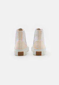 Sapatilhas altas de lona branca com acentos em bege e detalhes texturizados. Apresentam uma sola branca robusta e um logótipo castanho na parte de trás.