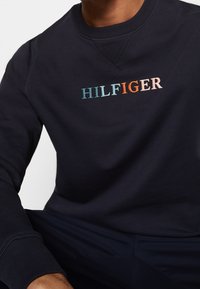 Mörkblå sweatshirt med rund halsringning, ribbade muddar och broderad "HILFIGER"-logotyp i aqua, orange och ljusrosa nyanser.