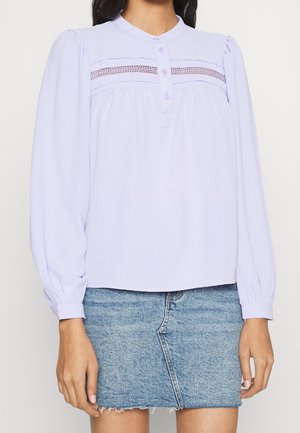 Femme portant un chemisier violet clair à manches longues avec des boutons et un détail en dentelle, associé à une mini-jupe en jean bleu avec un ourlet brut.
