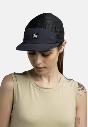 Femme portant une casquette en mesh noire avec le logo « Buff » et un haut beige sans manches, regardant vers le bas, montrant un tatouage sur le haut du bras.