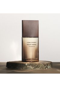 Bottiglia di profumo in vetro con bordi arrotondati, dotata di un tappo in legno scuro. Il liquido è di colore ambrato e viene presentato su una fetta di legno testurizzato.