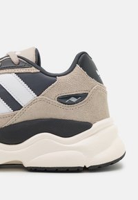 Sneaker beige e neri in materiale scamosciato e mesh, con strisce bianche e suola testurizzata. Accenti notevoli includono un dettaglio in plastica.