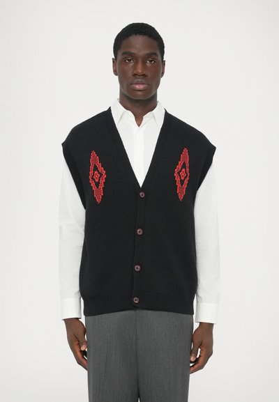 Joven vestido con un chaleco negro sin mangas abotonado con patrones geométricos rojos sobre una camisa blanca y pantalones grises, de pie frente a un fondo blanco.