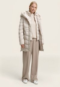 Beige gewatteerde jas met capuchon, ritssluiting en zijzakken. Draagt deze over een lichte trui en beige wijde broekspijpen, gecombineerd met witte sneakers.