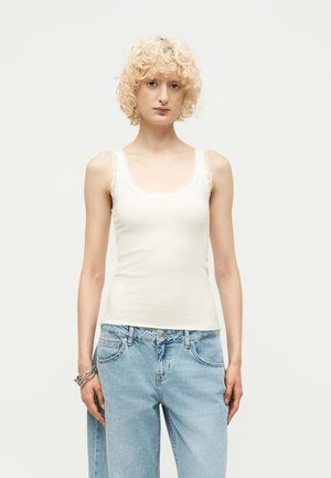 Jeune personne aux cheveux blonds courts et bouclés porte un débardeur blanc orné de dentelle et un jean taille haute bleu clair, debout devant un fond uni.