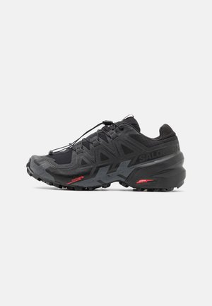 Black running shoe with synthetic upper, textured design, and contrasting red accents. Features a secured lace system and rugged outsole.