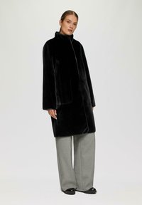Cappotto in pelliccia sintetica nera con colletto alto e vestibilità ampia. Presenta maniche lunghe e tasche laterali. Abbinato a pantaloni a gamba larga grigio chiaro.