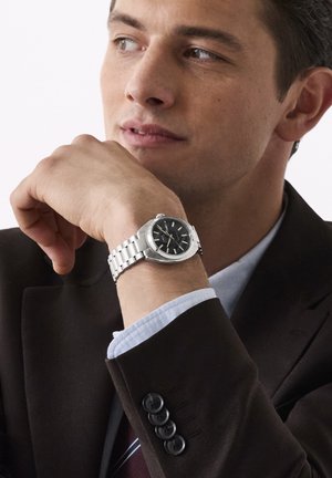 Homme en costume sombre posant le menton sur la main, portant une montre en métal argenté avec un cadran noir et un poignet de chemise bleu clair visible.