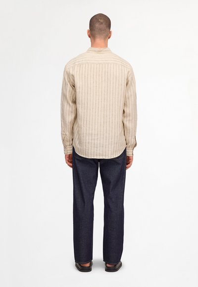 Man standing facing away, wearing a beige striped long-sleeve shirt, dark blue jeans, and black shoes against a white background.