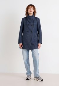 Navy double-breasted coat with a high collar, made of textured fabric, featuring six black buttons, paired with light blue jeans and sneakers.