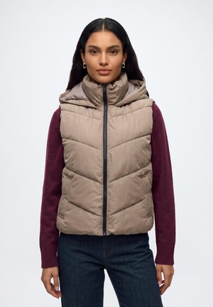 Femme aux longs cheveux foncés portant un gilet matelassé beige par-dessus un haut à manches longues bordeaux et un jean bleu foncé, debout devant un fond blanc uni.