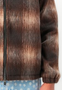 Bruine en beige fuzzy geblokte zip-up jack met elastische manchetten, gedragen over een blauwe stippenrok of -broek op een licht huid hand.