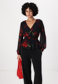 Blusa nera a fiori con stampa di rose rosse, scollatura a V, vita arricciata e maniche lunghe. Abbinata a pantaloni neri e una piccola borsa rossa.