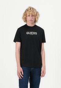 Juodas medvilnės marškinėliai su žodžiu "GUESS" raštuotame dizaine ir "EST. 1981" apačioje, trumpomis rankovėmis ir apvalia kaklo iškirpte.