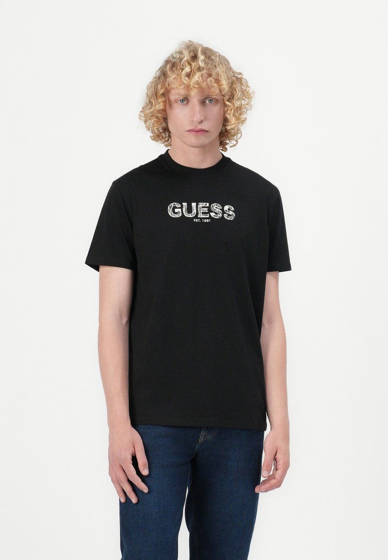 Juodas medvilnės marškinėliai su žodžiu "GUESS" raštuotame dizaine ir "EST. 1981" apačioje, trumpomis rankovėmis ir apvalia kaklo iškirpte.