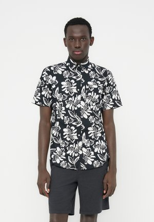 Hombre de pie con una camisa de manga corta con botones de flores en blanco y negro y pantalones cortos oscuros, sobre un fondo blanco liso.