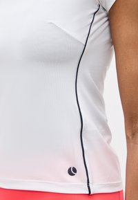 Κοντινό πλάνο ενός ατόμου που φοράει λευκό αθλητικό τοπ με μαύρο ρέλι κατά μήκος της πλαϊνής ραφής και ένα μικρό μαύρο λογότυπο κοντά στο τελείωμα.