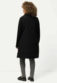 Musta tikattu pitkä liivi, jossa on timanttikuvio, yhdistettynä harmaisiin slim-fit-housuihin ja mustiin kenkiin. Liivissä on kaulus ja sivuvetoketju.