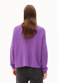 Eine Person mit langen braunen Haaren, die einen lockeren lila Pullover und schwarze Jeans trägt, von hinten vor einem weißen Hintergrund gezeigt.