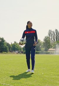 Adidas tepláková súprava v námorníckej modrej farbe s červeným akcentom, priliehavý strih. Drží čierno-biely futbalový loptu a stojí na zelenej tráve za denného svetla.