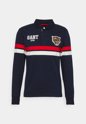 Polo de manga larga azul marino con rayas horizontales rojas y blancas, texto bordado GANT 1949 y parche con cabeza de tigre en el pecho.