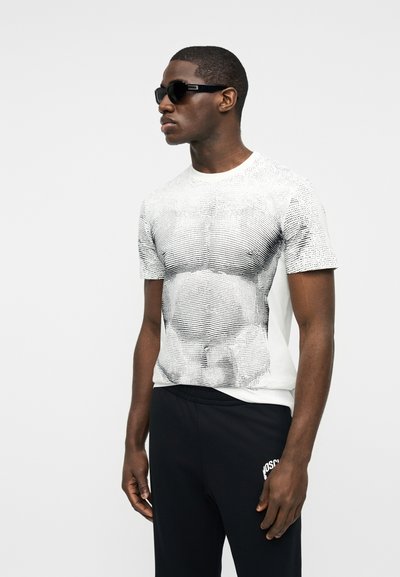 Άνδρας με μαύρα γυαλιά ηλίου, λευκό μπλουζάκι με μαύρες γραμμές περιγράμματος που μοιάζουν με μύες και μαύρο παντελόνι με λευκό κείμενο στον μηρό.