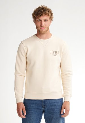 Man met krullend haar, gekleed in een crèmekleurige trui met het logo "PTRL Petrol Ind & Co." en een blauwe spijkerbroek, staand tegen een witte achtergrond.